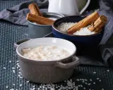 Gotowy do podania ryż na mleku w ceramicznej miseczce, obok miseczki z ryżem i laskami cynamonu.