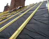 Charpente en bois et membrane noire sous une **ite toiture** en cours de construction, avec des cheminées en briques en arrière-plan.