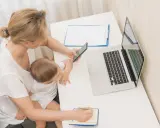 Kobieta z dzieckiem na kolanach sprawdza na telefonie, urlop macierzyński ile płatny. Obok laptop i notatnik.