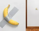 Un'opera d'arte banana, una gialla banana attaccata al muro con nastro adesivo grigio. A destra, un'altra banana, più piccola, fissata con nastro adesivo.