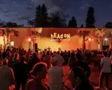 Crowd gathered at the hEAd oN photo festival, enjoying a performance under palm trees lit by warm orange lights.