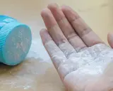Mano con borotalco bianco sparso, vicino al contenitore blu. Attenzione: il borotalco è cancerogeno.