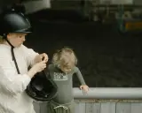 Starsza dziewczynka w kasku pokazuje młodszemu dziecku, jak zmierzyć głowę na kask, przygotowując się do jazdy konnej.