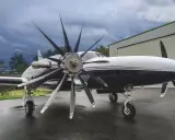 Ein elegantes Flugzeug mit einem beeindruckenden 4-Blatt-Propeller steht auf nassem Asphalt vor einer Halle.