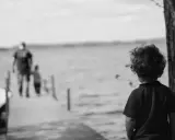 Bambino con capelli ricci guarda verso un molo sfocato dove camminano due figure. Un senso di isolamento, quasi un attaccamento disorganizzato, aleggia nell'aria.