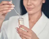 Une femme en peignoir blanc utilise un compte-gouttes pour prélever un sérum, peut-être un mélange de niacinamide et rétinol, pour sa routine de soins.