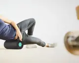 Frau rollt mit einer Faszienrolle über ihr Knie, um Muskelverspannungen zu lösen. Ein Skateboard ist im Vordergrund unscharf zu sehen.