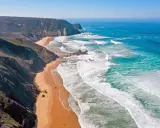 Atemberaubende Küstenlandschaft im Distrikt Faro, Portugal. Steile Klippen treffen auf einen goldenen Sandstrand und das türkisfarbene Meer mit tosenden Wellen.