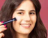 Una mujer sonríe mientras aprende como aplicar colorete con brocha, logrando un rubor natural en sus mejillas.