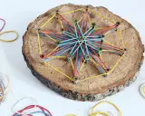 Bunte Gummibänder spannen sich zu einem Stern auf einer Baumscheibe. Ein tolles Bastelprojekt für Kinder, das Kreativität und Feinmotorik fördert.