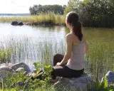 Kvinna sitter vid en sjö och utövar mindfulness svenska. Vassen vajar i vinden, solen glittrar på vattnet.