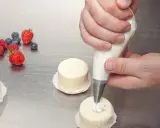 Man usa una manga pastelera para decorar pequeños pasteles. Frutas rojas y arándanos adornan la mesa.