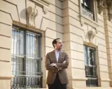 A man in a stylish brown sport coat outfit stands with arms crossed in front of a classic building.