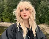 Blonde Frau mit lässigem Wolf Cut stylen, blauen Augen und einem schwarzen Jackett vor einer Waldkulisse.