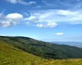 Zielone, trawiaste zbocze schodzi ku lasom i miastu w oddali. W tle widać kolejne pasma górskie i niebo z chmurami. To malownicze **beskidy szczyty**.