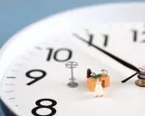 Figurka kobiety w sukience stoi przy miniaturowym biurku, symbolizując pracę w godzinach nadliczbowych. Zegar wskazuje późną godzinę.