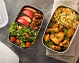 Dwa pojemniki z jedzeniem: jeden z kurczakiem w sosie, ryżem i natką, drugi z sałatką, jabłkami i orzechami. Idealne fit przepisy airfryer na lunch.