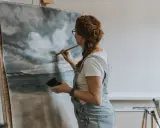 Konstnär med rött hår målar i olja en dramatisk himmel över ett hav.