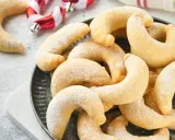 Ein Haufen goldbrauner Vanillekipferl, bestäubt mit Puderzucker, liegt auf einem dunklen Teller. Im Hintergrund sind weihnachtliche Zuckerstangen zu sehen.