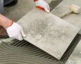 Układanie płytki na kleju w garażu. Ręce w białych rękawiczkach precyzyjnie kładą szarą płytkę na świeżym zaprawie.