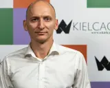 Łysy mężczyzna w białej koszuli na tle kolorowej ściany z logo "wKielcach".