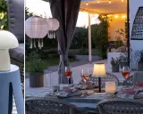 En mysig uteplats med dukat bord och flera lampor som skapar stämning. En vit svampformad lampa står på ett sidobord, medan papperslyktor och en flätad lampa hänger ovanför. Flera små bordslampor, liknande fotogenlampa utomhus, lyser upp bordet.