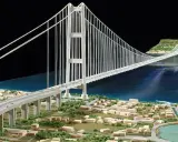 Maqueta de un puente colgante con pilares y cables que forman **puentes triangulados**, cruzando un cuerpo de agua y conectando dos orillas con una ciudad en miniatura.