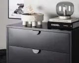 Svart sovrum med en mörk byrå. På byrån står en kruka med torkad växt, en lampa och böcker, bland annat "CHANEL CATWALK".