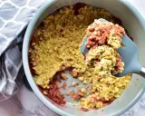 Ein köstlicher Erdbeer Rhabarber Crumble wird mit einem Löffel aus einer Auflaufform gehoben. Daneben liegt ein gemustertes Küchentuch.