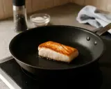 En bit torskfile steks gyllene i en stekpanna. Salt och pepparkvarn syns i bakgrunden.