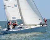 Un voilier blanc navigue sur une mer agitée, avec plusieurs personnes à bord. Le soleil brille, les voiles sont gonflées par le vent.