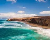 Fuerteventura w lutym: turkusowe fale rozbijają się o piaszczystą plażę, a za nią wznoszą się surowe, brązowe klify.