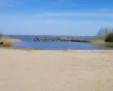 Plaża nad jeziorem Wicko, z pomostem, na którym stoją ludzie. W oddali widać trzciny i drzewa.