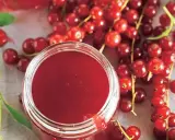 Ein Glas mit rotem Johannisbeergelee, umgeben von frischen Johannisbeeren. Perfekt zum Johannisbeeren einkochen.
