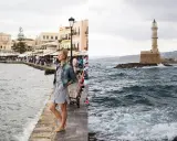 Kobieta spaceruje po promenadzie na Krecie. Październikowa pogoda, lekko zachmurzone niebo, morze faluje. W oddali widać latarnię morską.
