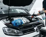 Persona con guantes rocía producto para limpiar motor coche. Usa cepillo para detallar el compartimento del motor cubierto de espuma.