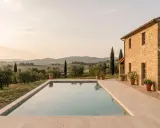 Piscina con vista sulle dolci colline pisane al tramonto. Una casa in pietra e ulivi completano il paesaggio idilliaco.
