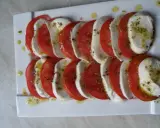 Mozzarella z pomidorami, ułożona naprzemiennie na białym talerzu, skropiona oliwą i ziołami.