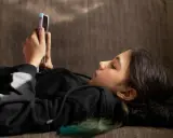 Ein Mädchen liegt auf einer Couch und schaut auf ihr Smartphone. Sie ist in ein schwarzes Kapuzensweatshirt gekleidet. Dies spiegelt aktuelle Trends der Jugend wider.