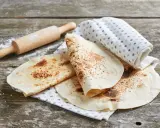 Świeżo usmażona tortilla z patelni, lekko przypieczona, leży na lnianej ściereczce obok wałka.