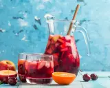En karaff med röd bål, fylld med frukt och is, står bredvid två glas. Äpplen, körsbär och en apelsinskiva dekorerar bordet. Ett perfekt bål recept för sommaren.
