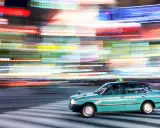 Un taxi azzurro sfreccia su strisce pedonali, con luci sfocate di città che creano un effetto dinamico, perfetto per imparare come fare foto in movimento.