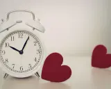 Biały budzik pokazuje 10:08. Obok niego leżą dwa czerwone serca. 0505 znaczenie - czas na miłość.