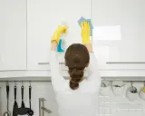 Mujer con guantes amarillos realiza limpieza armarios cocina, rociando y limpiando con paño azul.