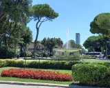 Fontana z wodą w **Parco Fellini Rimini**, otoczona zielenią i kwiatami. W tle widać autobus i wieżowiec.