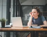 Zmęczona dziewczyna przy biurku, zamyślona, jakby zastanawiała się, ostracyzm co to. Laptop, książki i roślina na blacie.