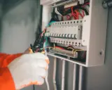Elektryk w białych rękawicach manipuluje przewodami w skrzynce z bezpiecznikami. Wybija bezpiecznik bez obciążenia, co może sugerować problem.