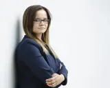 Kobieta w okularach i granatowej marynarce stoi oparta o ścianę, ze skrzyżowanymi rękami.