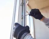 Ręce w rękawiczkach dokonują regulacji drzwi zewnętrznych PCV, dokręcając śrubę kluczem.