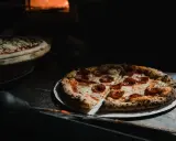 Upieczona pizza pepperoni na talerzu, obok surowa pizza w piecu.